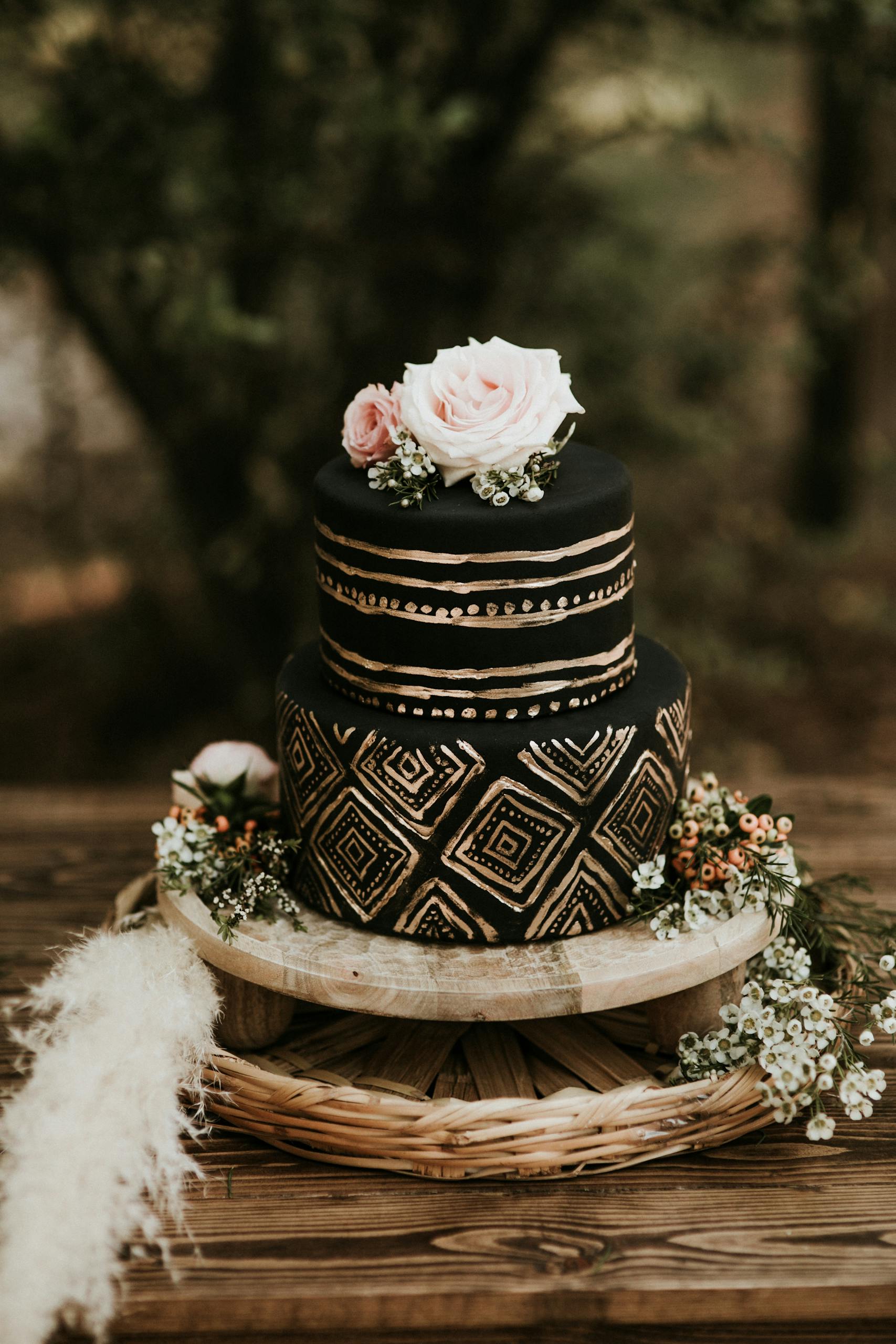 A stunning black and gold wedding cake with floral decorations on a wooden table outdoors.